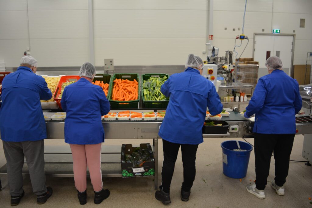 Elintarvikelinjasto, jossa pakataan keittovihanneksia styroksirasioihin. Linjaston edustalla sinisiin työtakkeihin pukeutuneita työntekijöitä.